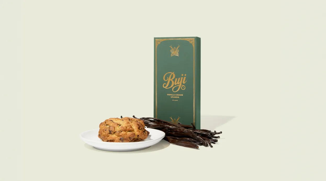 Box of Buji Uganda vanilla beans with vanilla pods and a cookie on a white plate.