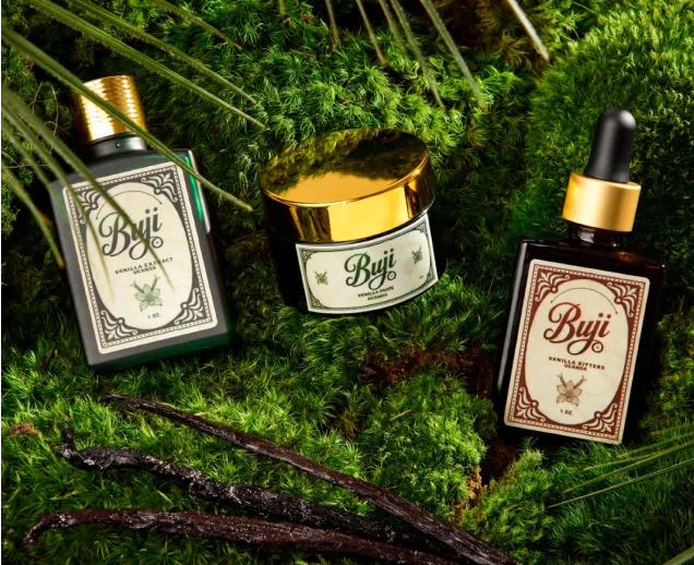 Three Buji vanilla extract products on green moss surrounded by vanilla pods and plant leaves.