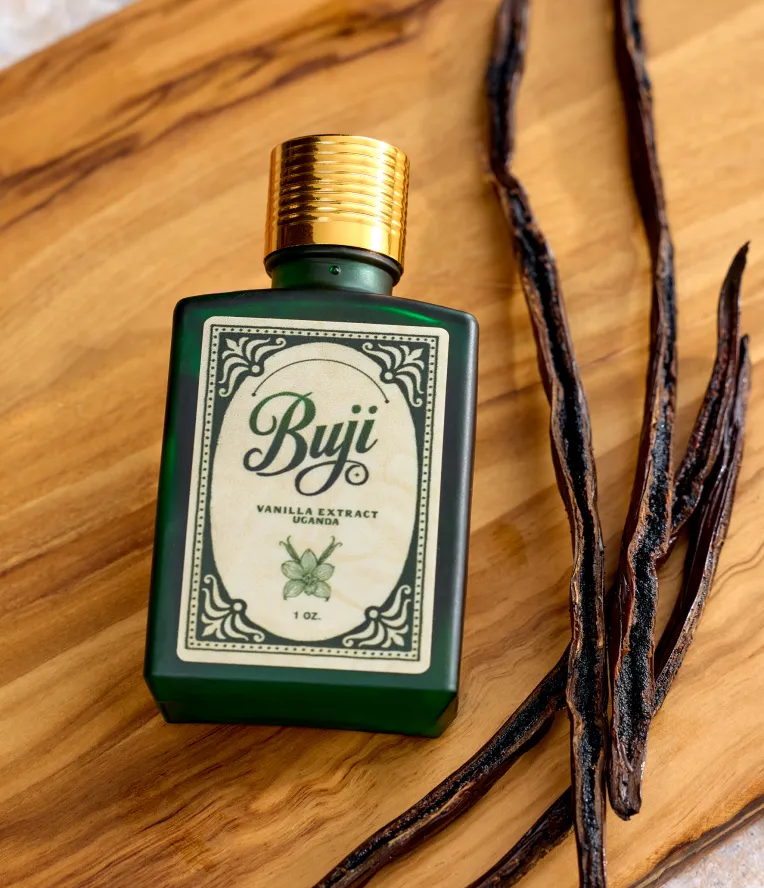 Dark green bottle of Buji vanilla extract with golden cap beside dried vanilla pods on wood surface.