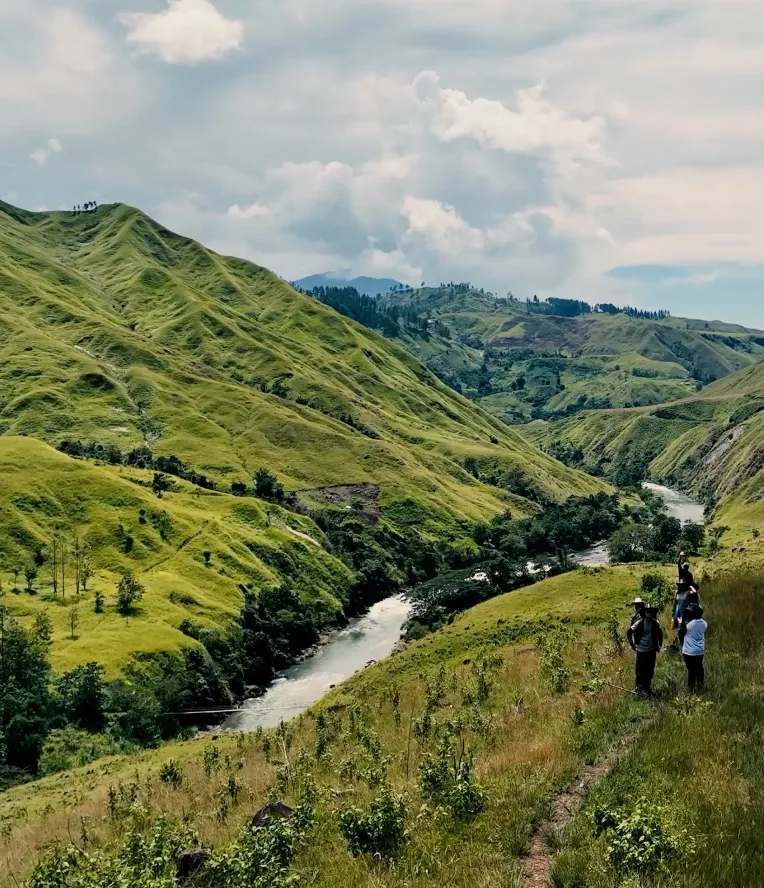 People standing on a grassy hill overlooking a river winding through green, rolling hills under a partly cloudy sky.