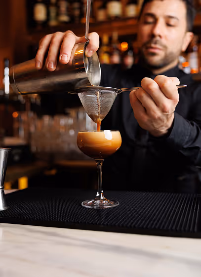 Bartender double-straining a foamy cocktail into a coupe glass at the bar