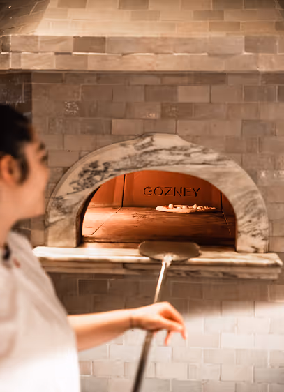 Chef sliding a pizza into a marble-fronted stone oven on a wooden peel