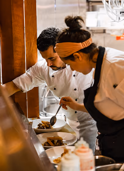 Two chefs in whites plating dishes together at the pass in a busy kitchen