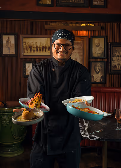 Chef balancing four Indian dishes beneath framed vintage portraits in a red-boothed dining room