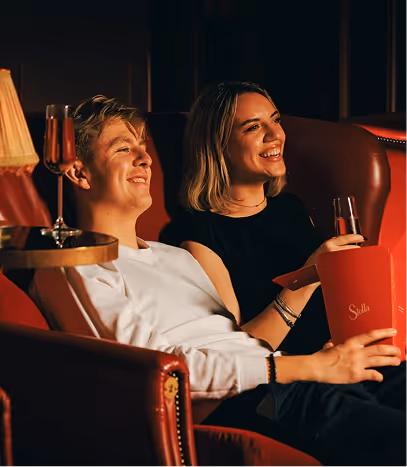 Guests laughing with cocktails and a Stella popcorn tub in the red armchairs at Stella Cinema