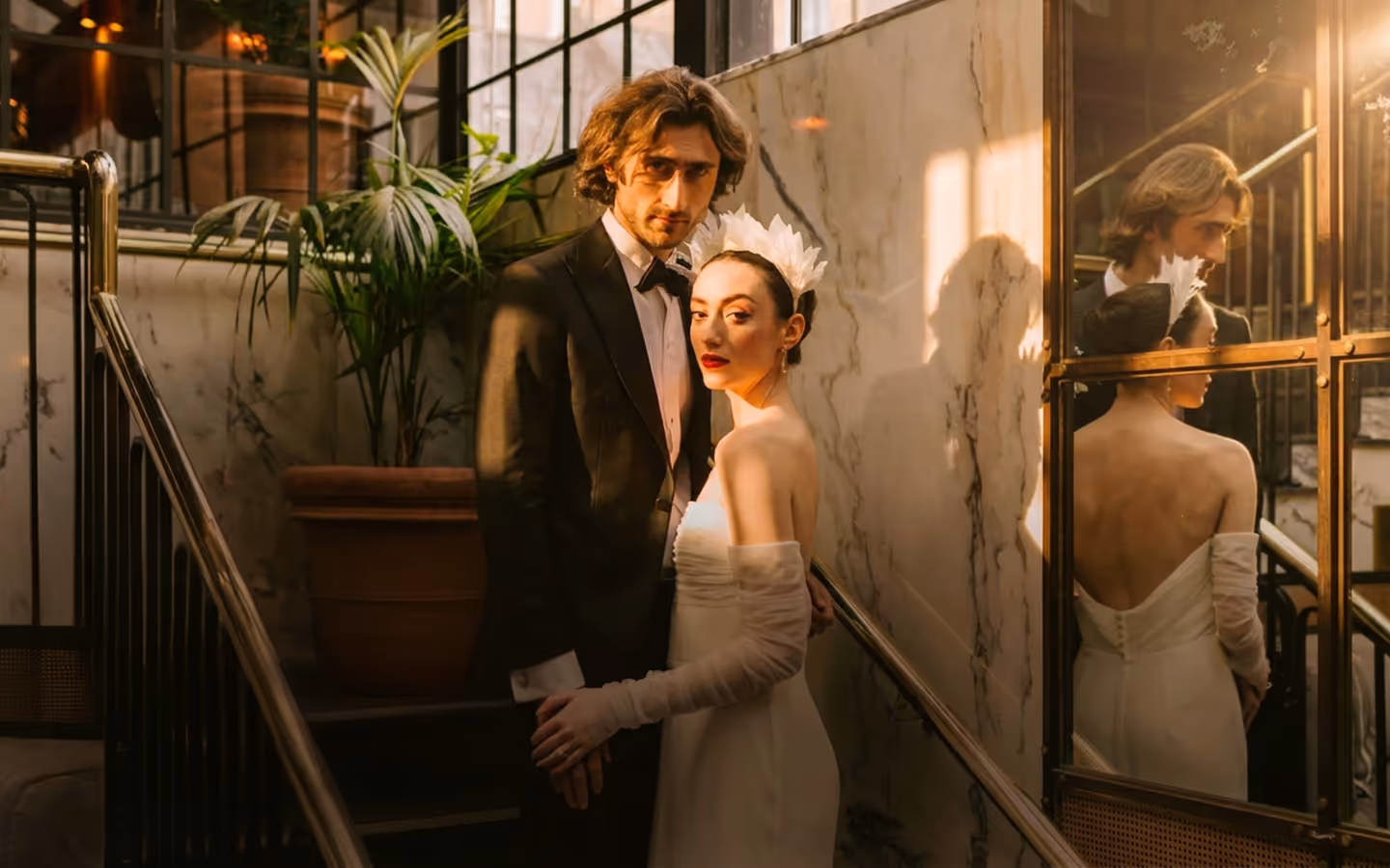 Bride in a feathered headpiece and groom in black tie on a marble staircase, mirrored behind them