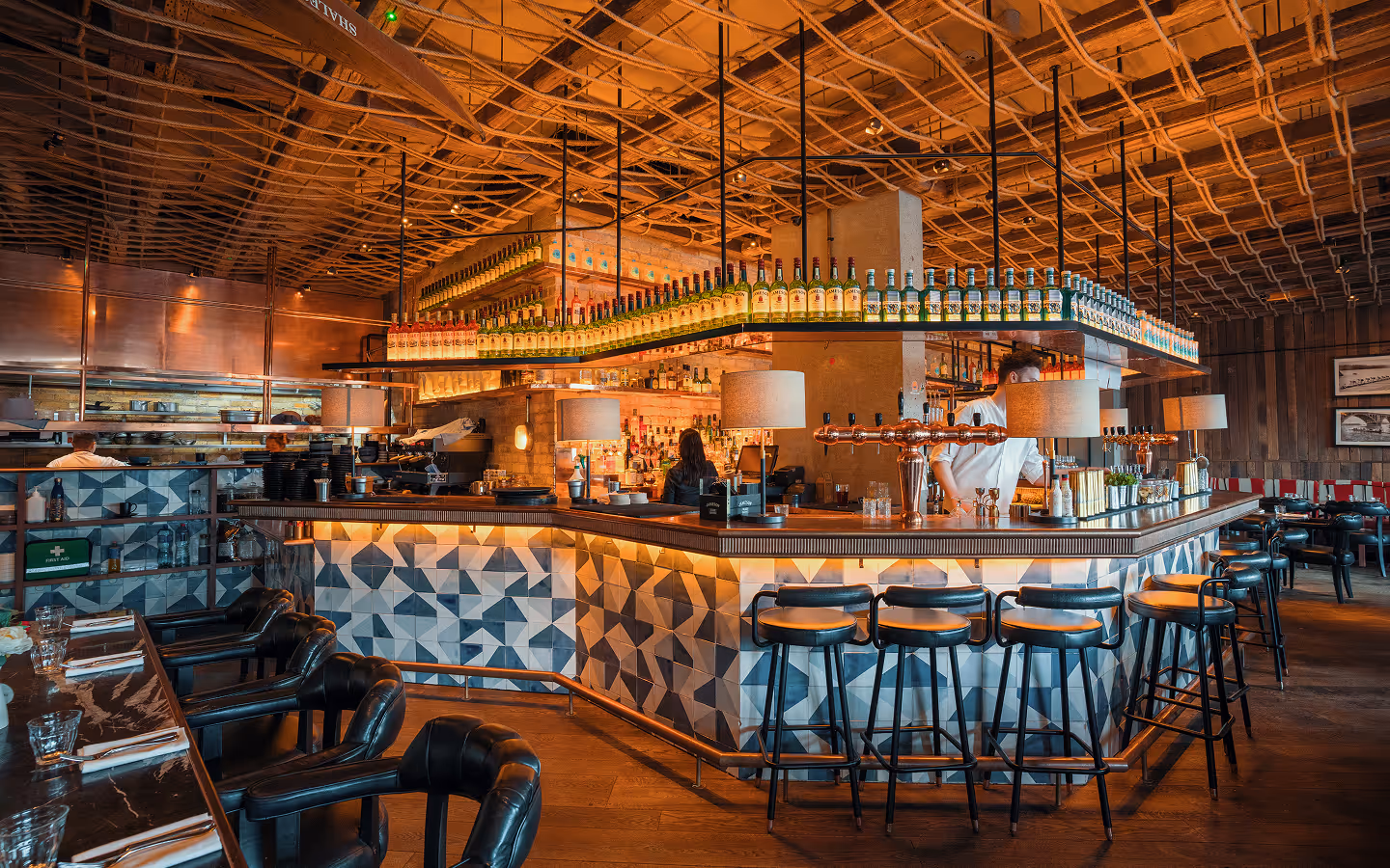 Tiled island bar under a rope-lattice ceiling, with bar stools and leather tub chairs in the dining room