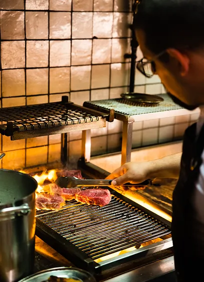 Chef laying steaks onto an open charcoal grill in a tiled kitchen