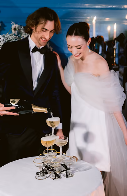 Groom pouring champagne into a coupe tower as the bride watches at a candlelit reception