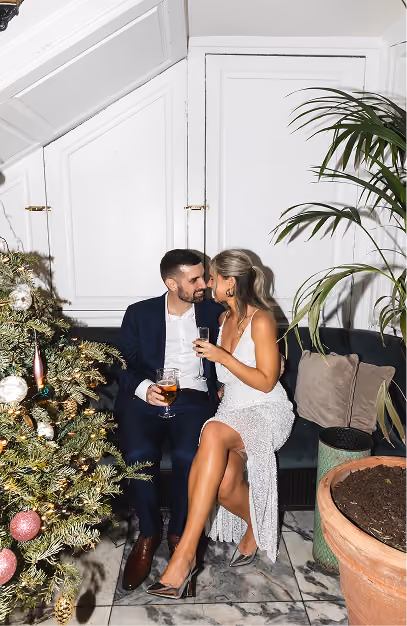 Couple in evening dress sharing drinks on a green banquette beside a decorated Christmas tree