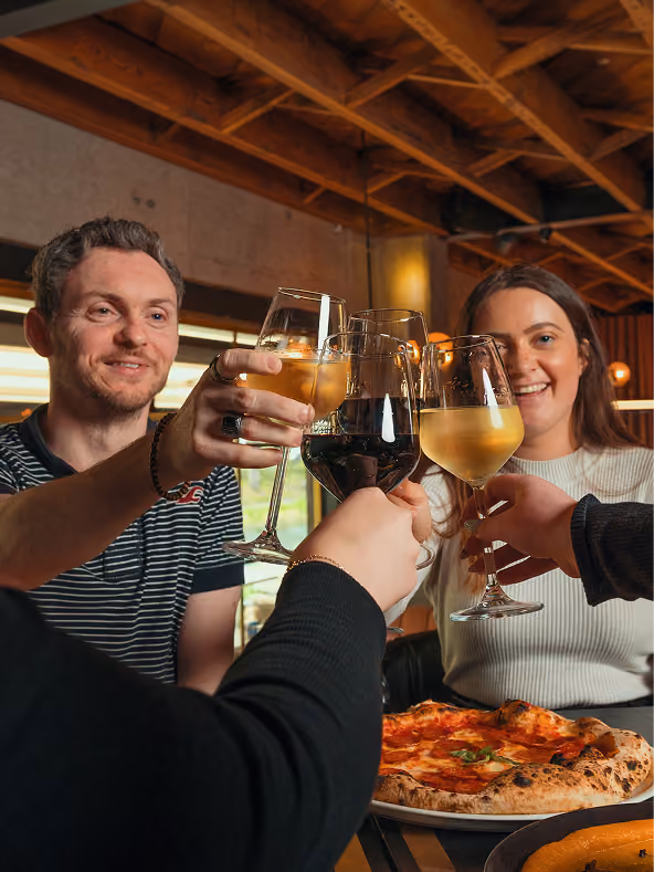 Friends clinking wine glasses over a freshly baked margherita pizza at a restaurant table