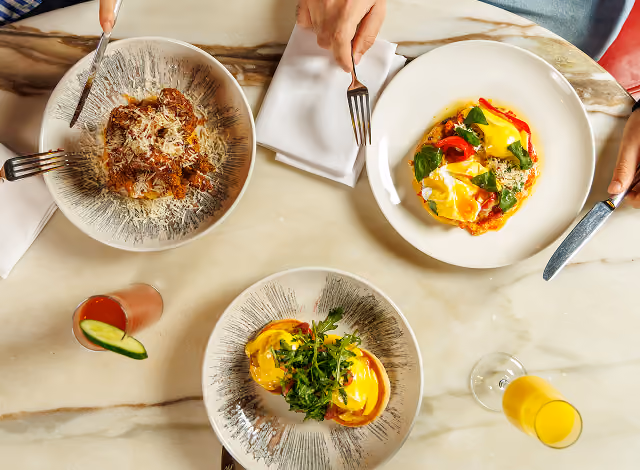 Overhead brunch spread on marble: eggs with hollandaise, pasta and a cocktail