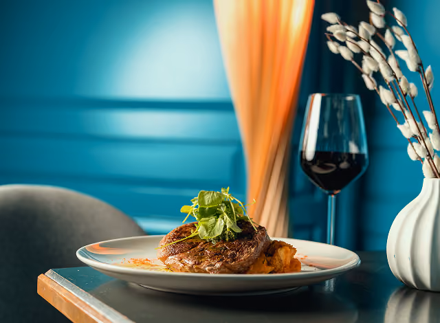 Seared steak topped with herb salad beside a glass of red wine against a teal panelled wall