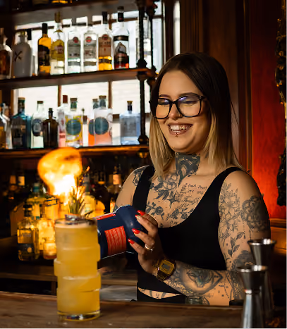 Tattooed bartender holding a shaker beside a garnished cocktail at the bar