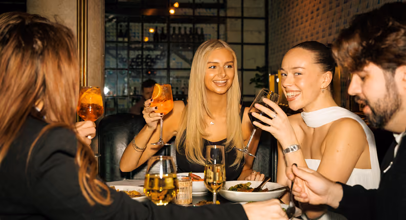 Friends raising spritzes and cocktails over dinner in a low-lit restaurant