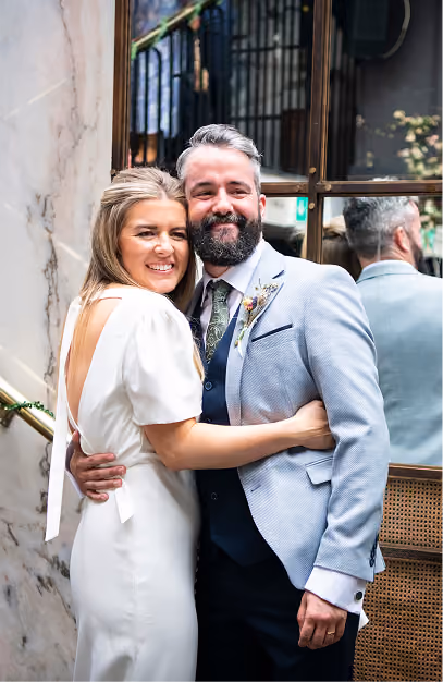 Newlywed couple embracing against a marble wall, groom in a pale grey suit with buttonhole