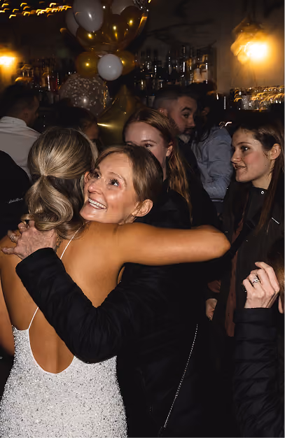 Bride in a sequinned dress hugging a guest at a busy bar under gold and white balloons