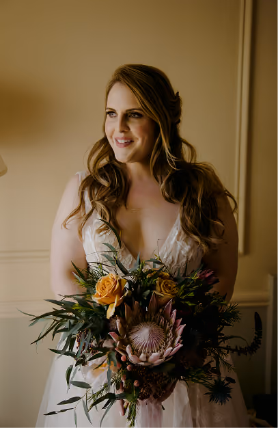 Bride holding a bouquet of king protea, orange roses and eucalyptus in a cream-panelled room