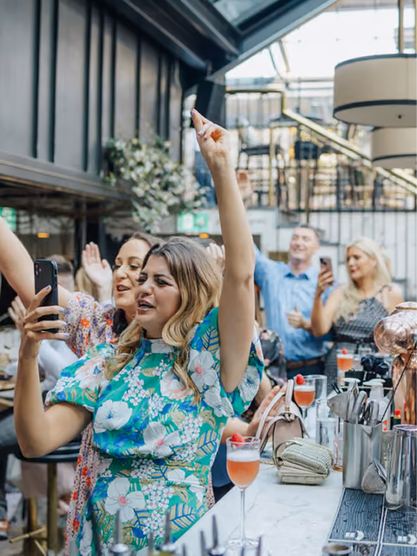 Guests cheering and filming on a phone at a marble bar, cocktails lined up on the counter