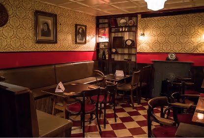 Cozy vintage pub interior with patterned wallpaper, framed portraits, cushioned bench seating, wooden chairs and tables, checkered floor, and a dark fireplace.