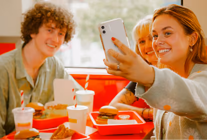 Three smiling young adults sitting at a fast-food table with burgers, fries, and drinks, as one woman takes a selfie with a smartphone.