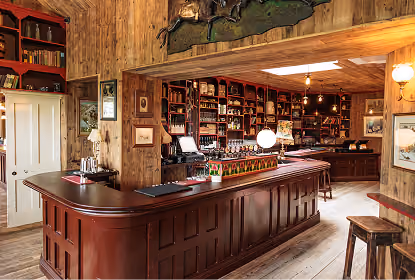 Interior of a rustic wooden pub with a long dark wood bar counter, shelves filled with bottles and glasses, wooden stools, and soft warm lighting, creating a cozy atmosphere.