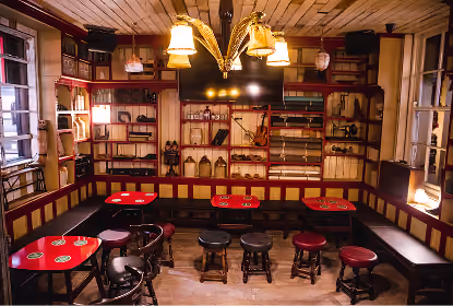 Cozy pub interior with wooden walls and ceiling, cushioned bench seating around the room, red tables, wooden stools, shelves with bottles and decorations, and a large TV mounted on the back wall under hanging lamps.