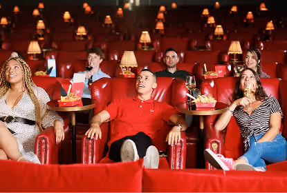 People seated in comfortable red leather chairs in a dimly lit cinema, watching a movie, with popcorn and drinks on trays attached to the chairs.