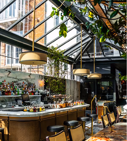 Bright modern bar interior with a high glass ceiling, hanging pendant lights, a long marble counter with black bar stools, bottles and glasses behind the bar, and lush green plants around.