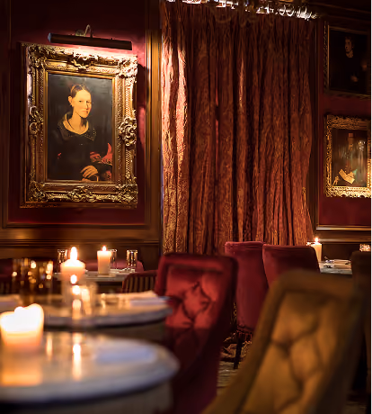Cosy, dimly lit room with red velvet chairs, round tables with lit candles, and classic framed paintings on walls, creating an intimate atmosphere.
