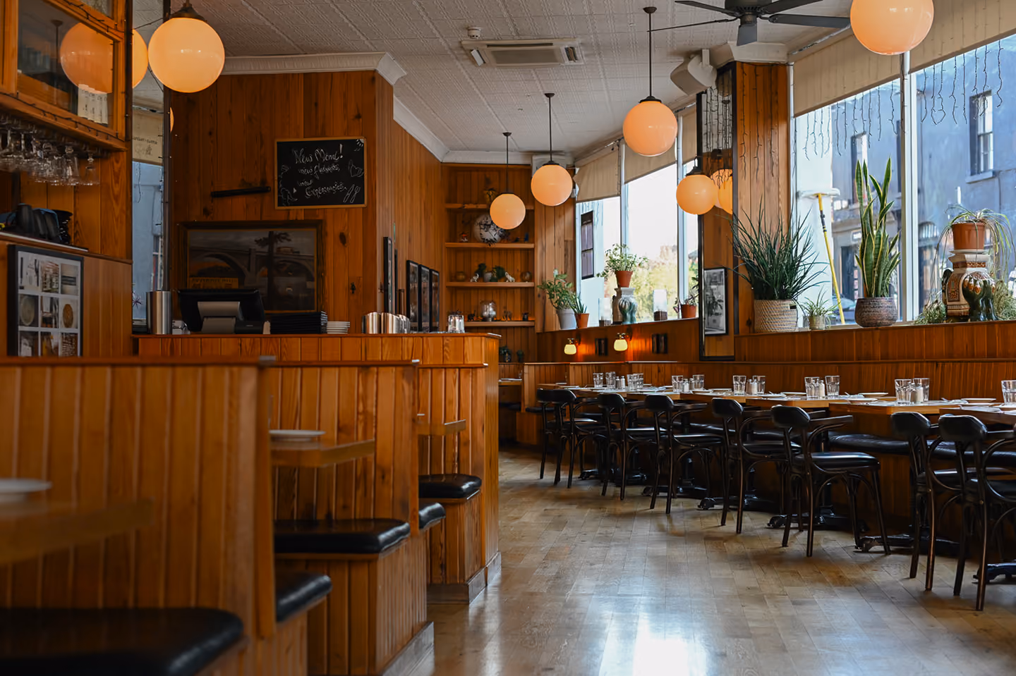 Interior of a cosy restaurant featuring wooden booth seating with black cushions on the left and a row of tables with black chairs set with glasses and utensils on the right, large windows with plants on the ledges, and round pendant lights hanging from the ceiling.