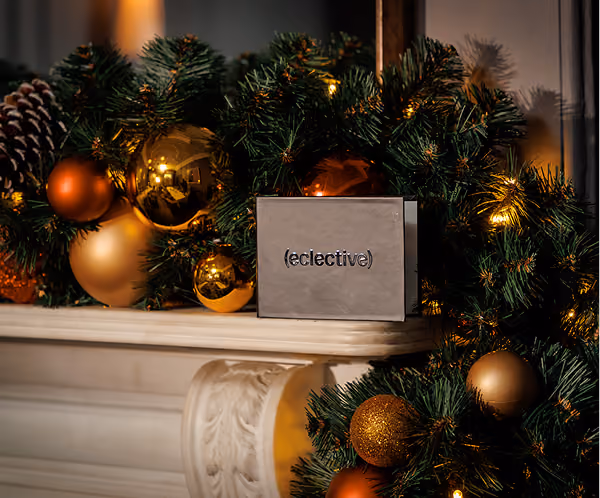 Festive Christmas mantel decorated with green pine garland, warm white string lights, and shiny gold and bronze baubles, with a small rectangular card reading '(eclective)' at the center.