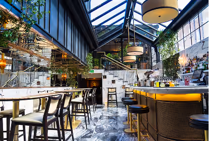 Bright, modern bar interior with marble flooring, high ceiling with glass panels, illuminated bar counter with bar stools, tables and chairs, and hanging lamps.