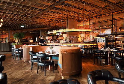 Interior of a stylish restaurant with wooden floors and ceiling, black leather chairs, wooden tables, a bar stocked with bottles, and warm ambient lighting.