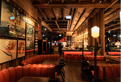 Cozy, warmly lit bar interior featuring brown leather booth seating, wooden walls with art frames, a shelf with bottles, a bar counter with stools, and a lit floor lamp.
