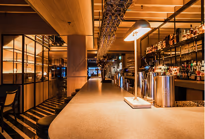 Interior view of a modern bar with a long beige countertop, bar stools, warm lighting, glass racks hanging overhead, and shelves stocked with bottles of liquor in the background.