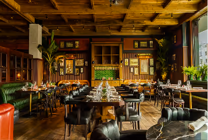 Interior of a stylish restaurant with wooden ceiling, wood-paneled walls, and a variety of seating including green leather booths and black cushioned chairs around neatly set tables.