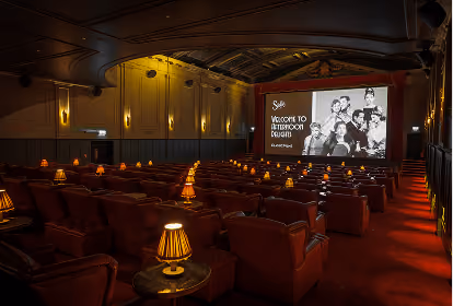 Interior of a dimly lit vintage cinema with rows of red armchairs and small lamps on side tables, facing a screen displaying a black and white photo and text welcoming to the Jefferson Deluxe cinema.