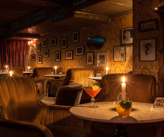 Dimly lit lounge with brown velvet armchairs and round marble tables, each table holding a lit candle and drinks, and walls decorated with framed pictures.
