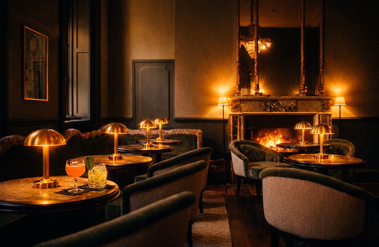 Cosy lounge with multiple round tables, upholstered chairs, and vintage-style golden lamps, centered around a lit fireplace with a large mirror and two wall lamps.