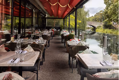 Outdoor restaurant seating area with tables and chairs arranged neatly along a canal, featuring glass railings, red canopy overhead, and a stone bridge visible in the background.