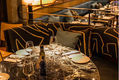 Round marble table set for four with clear wine glasses, plates, cutlery, and a bottle of olive oil and pepper grinder, surrounded by a cushioned bench with dark fabric and gold geometric patterns in a dimly lit restaurant.