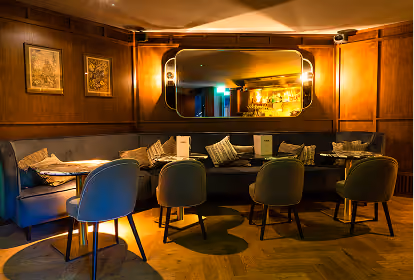 Cozy lounge area with dark wood panel walls, a large horizontal mirror, blue and green upholstered chairs, a banquette with cushions, and soft warm lighting.