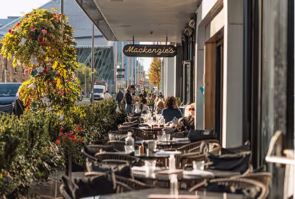 Outdoor seating area of a restaurant called Mackenzie's with tables, chairs, and people dining along a sidewalk under a gray awning in a city setting with greenery and a large floral arrangement.