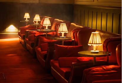 Row of red velvet armchairs with round side tables each holding a small lamp with pleated shades, arranged against a wall in a dimly lit, cozy room.