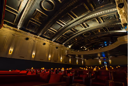 Interior of a classic movie theater with red velvet seats, warm wall sconces, and an ornate decorative ceiling with gold accents.