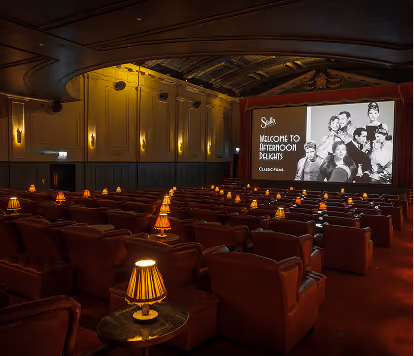 Classic cinema-style venue featuring rows of red leather seats, warm table lamps, and a large movie screen.