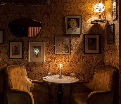 Intimate vintage-style seating area with two armchairs, a round table, candlelight, and ornate patterned wallpaper.