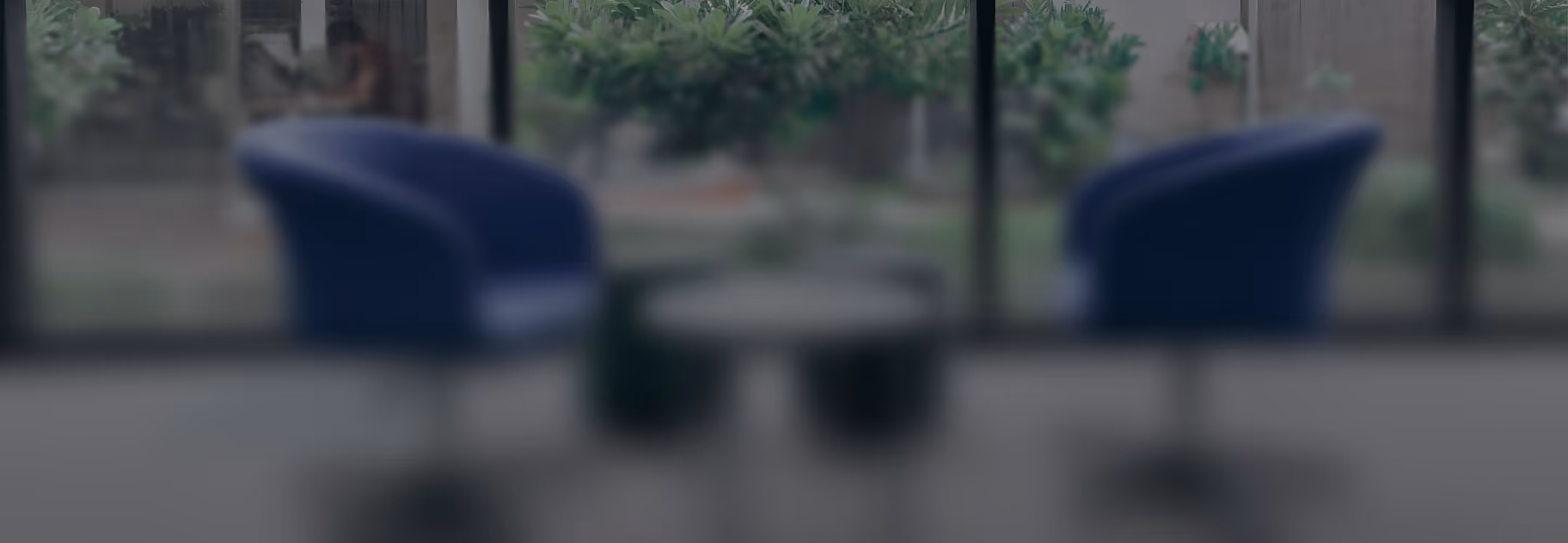 Two blue chairs facing each other with a small round table in front of glass windows and plants.