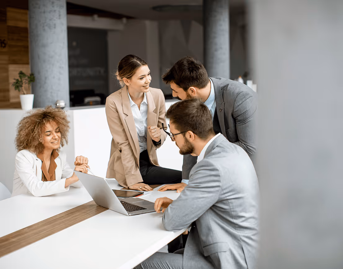 Four businesspeople collaborating around a laptop at a white conference table in an office.